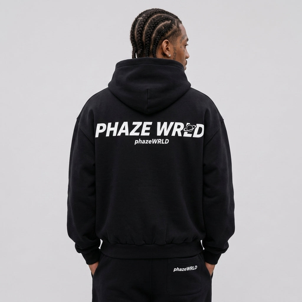 Rear view of a model wearing a premium black oversized streetwear hoodie with a bold white "Phaze Wrld" planet graphic print on the back.