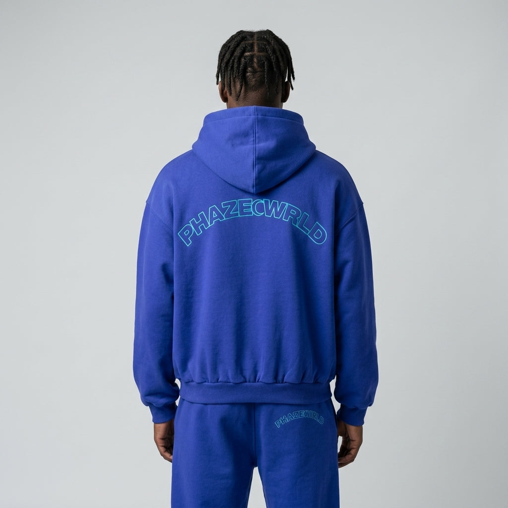 Back view of a premium cobalt blue oversized streetwear hoodie with a teal arched 'PHAZEWRLD' outline graphic on the shoulders.