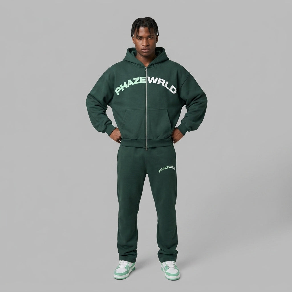 A studio lookbook photo of a streetwear model wearing a premium dark green oversized zip-up hoodie with an arched chest graphic.