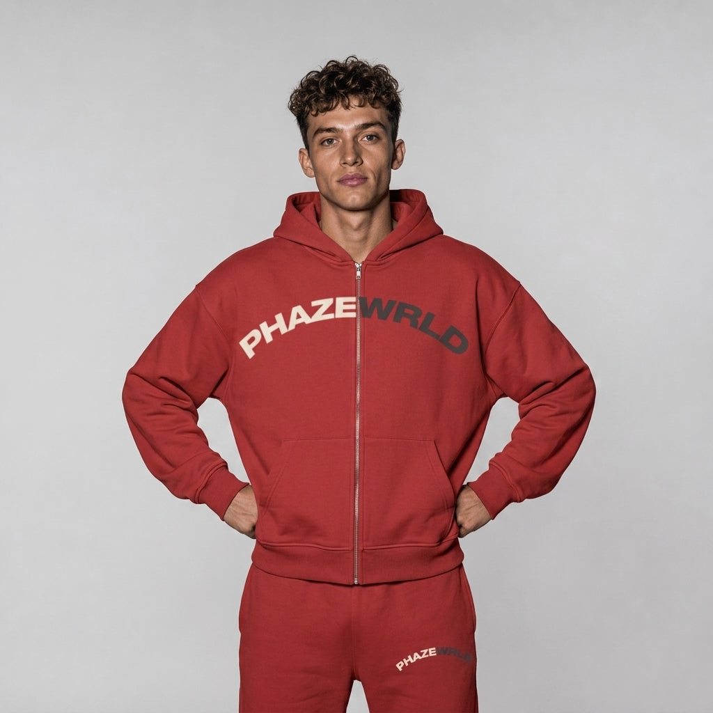 An e-commerce lookbook photo of an oversized crimson red full-zip streetwear hoodie with a cream and charcoal arched "PHAZE WRLD" graphic across the chest.