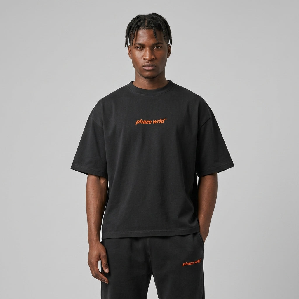 An oversized washed black heavyweight streetwear t-shirt featuring a small vibrant orange "phaze wrld" logo centered on the chest.