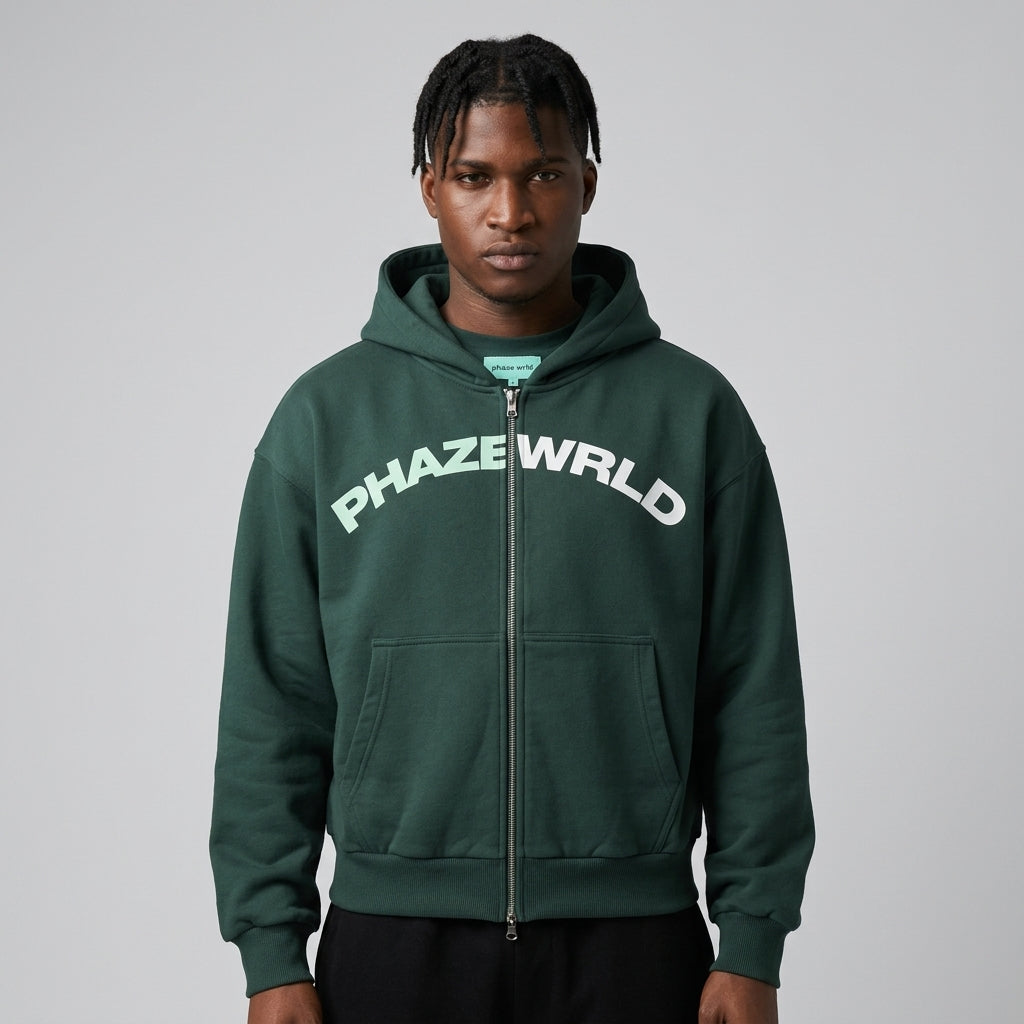 A studio lookbook photo of an oversized pine green streetwear zip-up hoodie with an arched mint and white PHAZEWRLD logo across the chest.